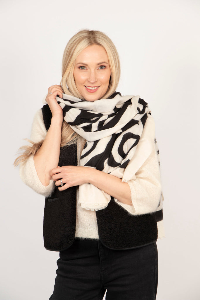 model wearing a swirling pattern cream and black lightweight scarf wrapped around the neck and shoulders.