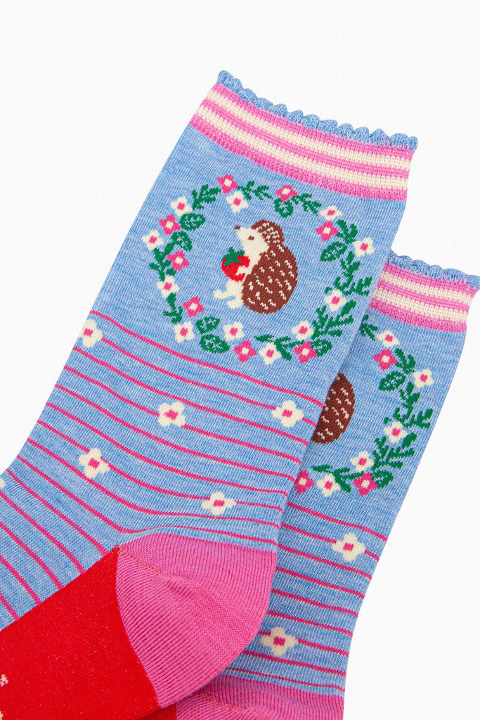 close up of the pattern. on the ankle of the socks is a large brown and beige hedgehog standing on its hind legs. it is holding a red strawberry in its hands. surrounding the hedgehog is a floral wreath made of white flowers, pink flowers and green leaves. 