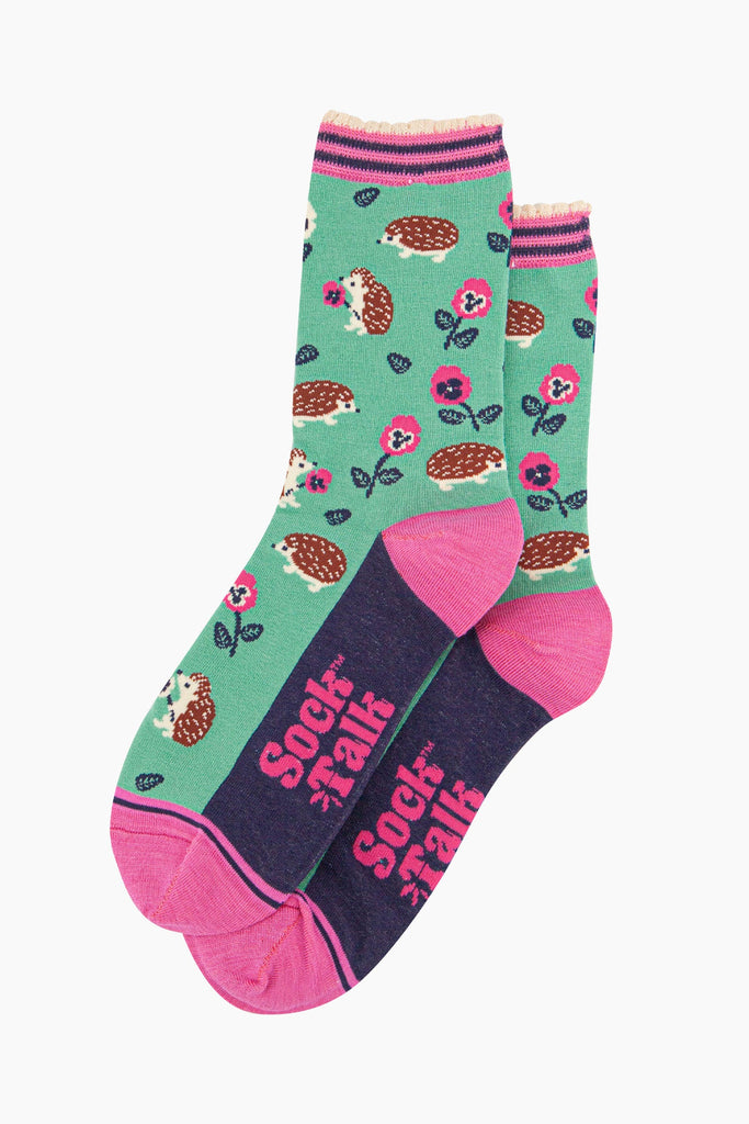 the pair of mint green hedgehog socks laying flat one overlapping the other. showing clearly the green background of the socks which is contrasted by the all over pattern of hedgehogs, flowers and leaves.  the sock talk logo is pink and visible on the base of the sole. 