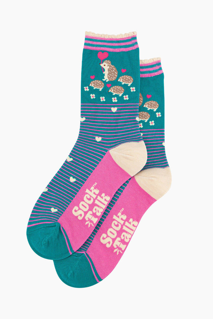 the pair of teal hedgehog socks laying flat one overlapping the other. showing clearly the teal background of the socks which is contrasted by pink pinstripes. on the ankle of the socks there is a woodland scene with a family of hedgehogs.  the sock talk logo is visible on the base of the sole. 