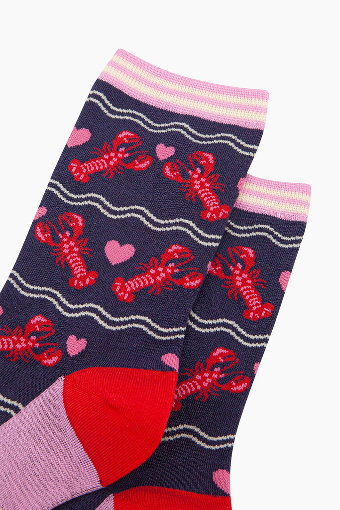 close up of the pattern, the lobsters are red with pink highlights on their backs and pincers, they are in pairs with a pink love heart between them. the cuff of the socks is straight cut and is light pink and cream striped.