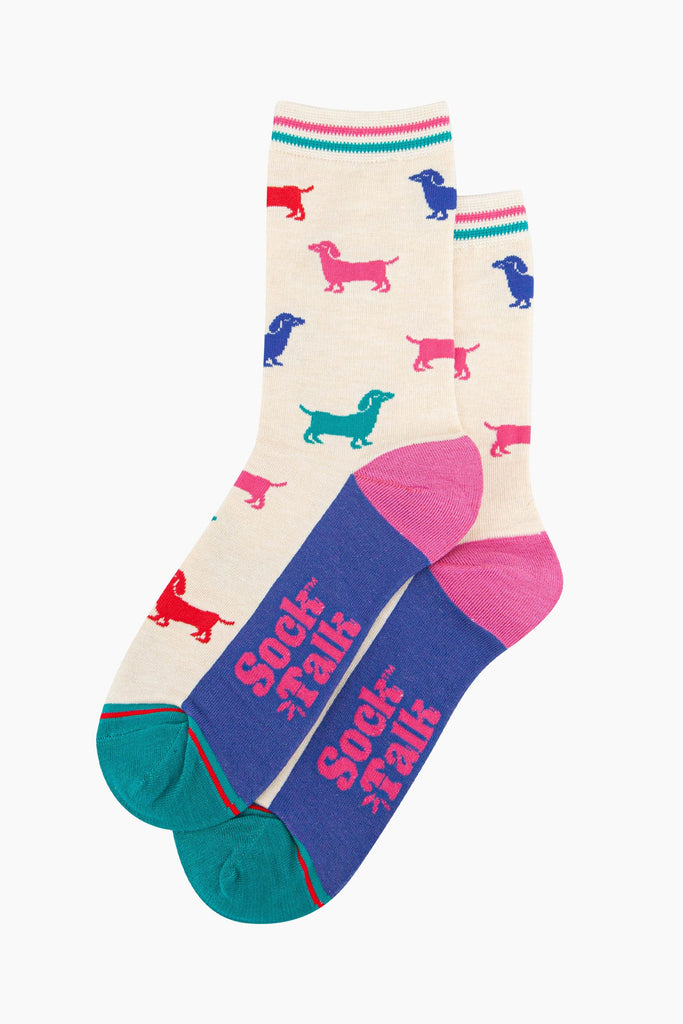 the pair of colourful sausage dog socks laying flat, one overlapping the other. showing clearly the cream background of the socks which is contrasted by colourful sausage dog silhouettes. the sock talk logo is visible on the base of the sole. 