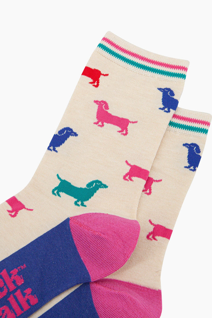 close up of the pattern. the sausage dogs are a variety of bold colours, royal blue, turquoise, pink and red. the cuff has a pink and a turquoise horizontal stripe which contrasts the cream background of the socks.