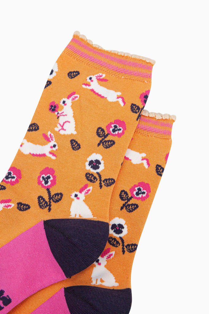 close up of the pattern, the white rabbits are running, sitting and jumping, some are holding flowers. the floral pattern features pink and white pansy like flowers. the cuff is scalloped and has a pastel orange and pink striped pattern.