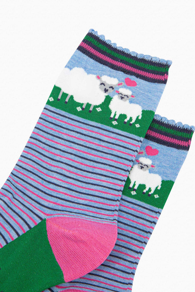 close up of the pattern, the ankle part of the socks features a white sheep and a baby lamb. between the animals is a pink love heart. the cuff of the socks is scalloped and has a pink and green stiped pattern.