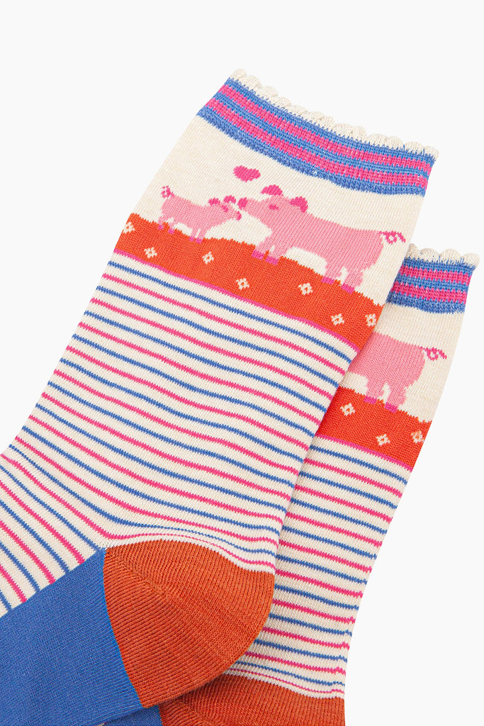 close up of the pattern, the ankle part of the socks features a pink mother pig nuzzling her baby piglet. between the pigs is a pink love heart. the cuff of the socks is scalloped and has a pink and blue stiped pattern.