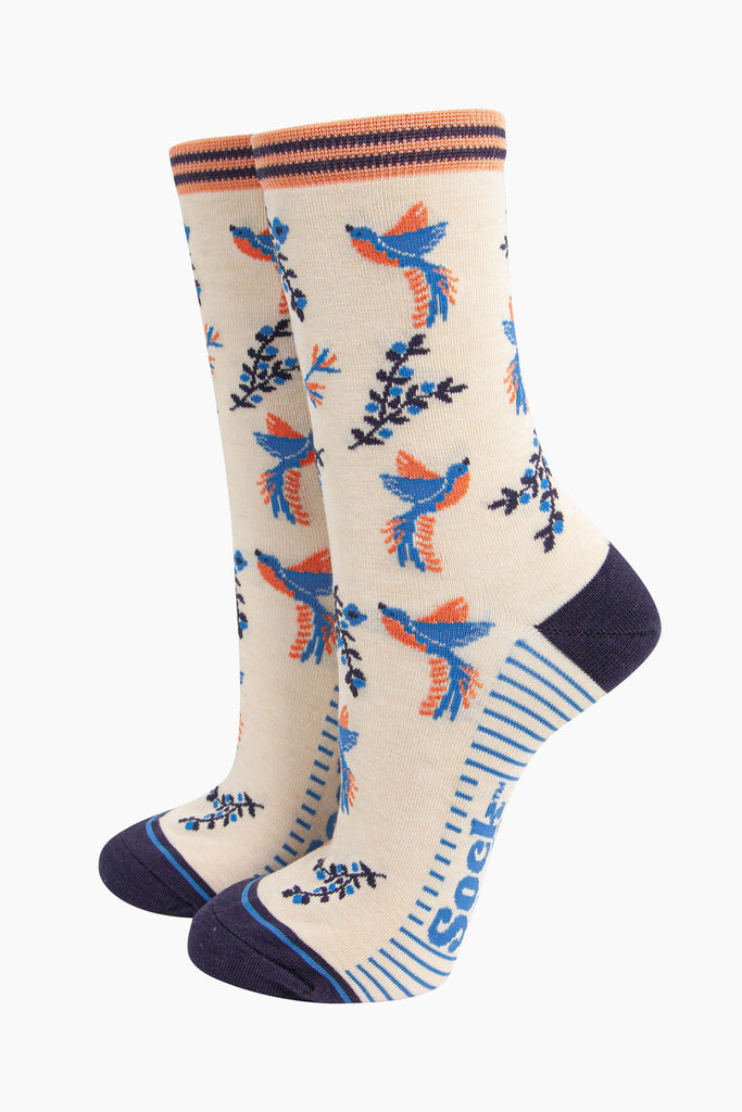 cream hummingbird floral bamboo socks. the hummingbirds and blue and orange and are shown in flight, hovering around floral vines. the toe and heel of the socks are a contrasting navy blue.