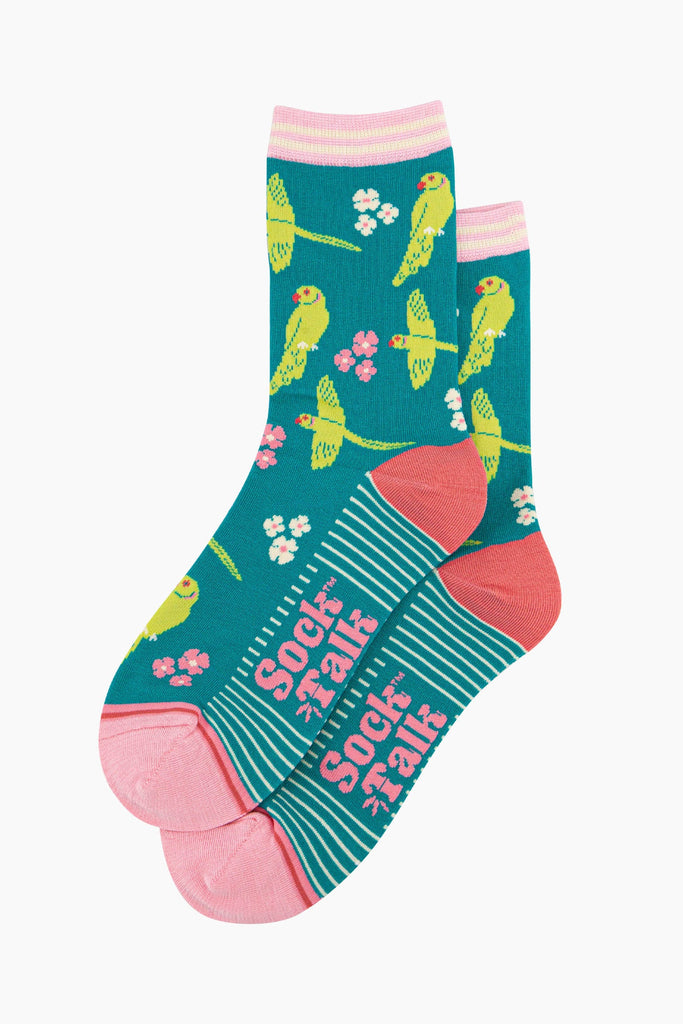 the pair of teal parakeet socks laying flat, one overlapping the other. showing clearly the teal blue background of the socks which is contrasted by the lime green parakeets and white and pink flowers. the sock talk logo is visible on the base of the sole. 