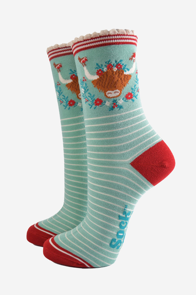 mint green bamboo socks with a large highland cow on the ankle, the highland cow is surrounded by red robin birds, and holly, the socks have a scalloped edge and a white pinstripe