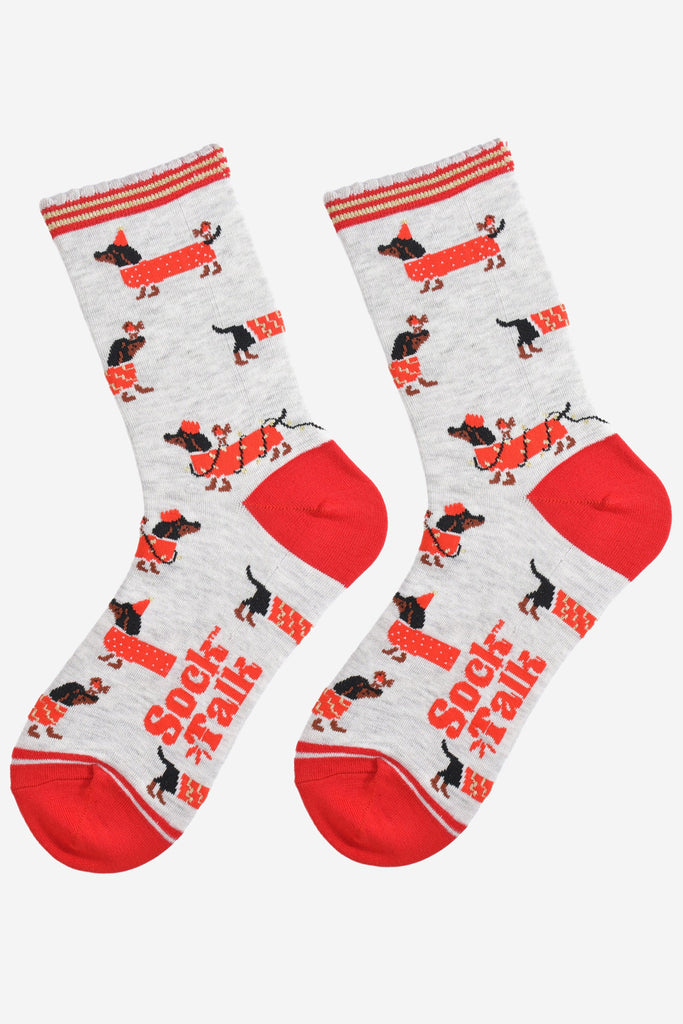 showing the socks laying flat, there is an all over pattern of black sausage dogs wearing red festive coats, party lights and hats. there are also red robins playing with the sausage dogs