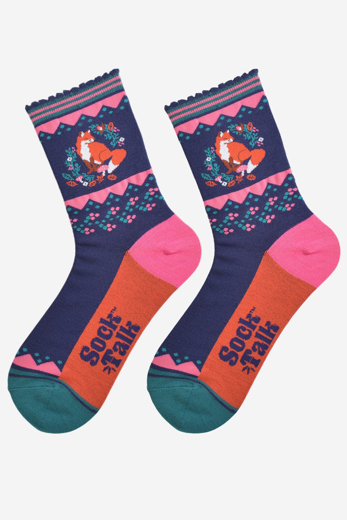 fox and wreath socks laying flat, showing the orange sole, pink heel and green toe which contrast the navy blue socks, on the ankle of the socks ther is a red fox enclosed in an wreath of autumnal foliage