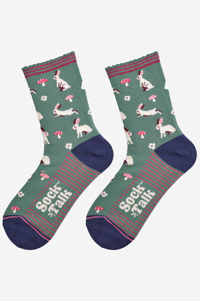 showing the woodland rabbit socks laying flat, there are white rabbits, hopping, sitting and standing among pink toadstools and white flowers, the solve of the socks has pink stripes in contrast to the green base of the design