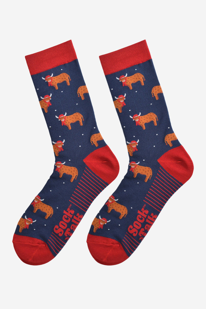 navy blue and red highland cow bamboo socks laying flat, the highland cows are shown in profile and they are standing up, they are wearing red santa hats and red winter scarves