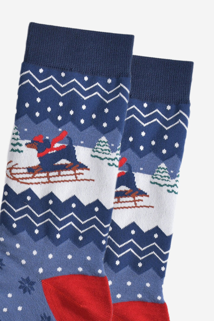 close up of the sledging sausage dog on the ankle of the socks, the dog is wearing a red hat and scarf and there are snow covered christmas trees in the background