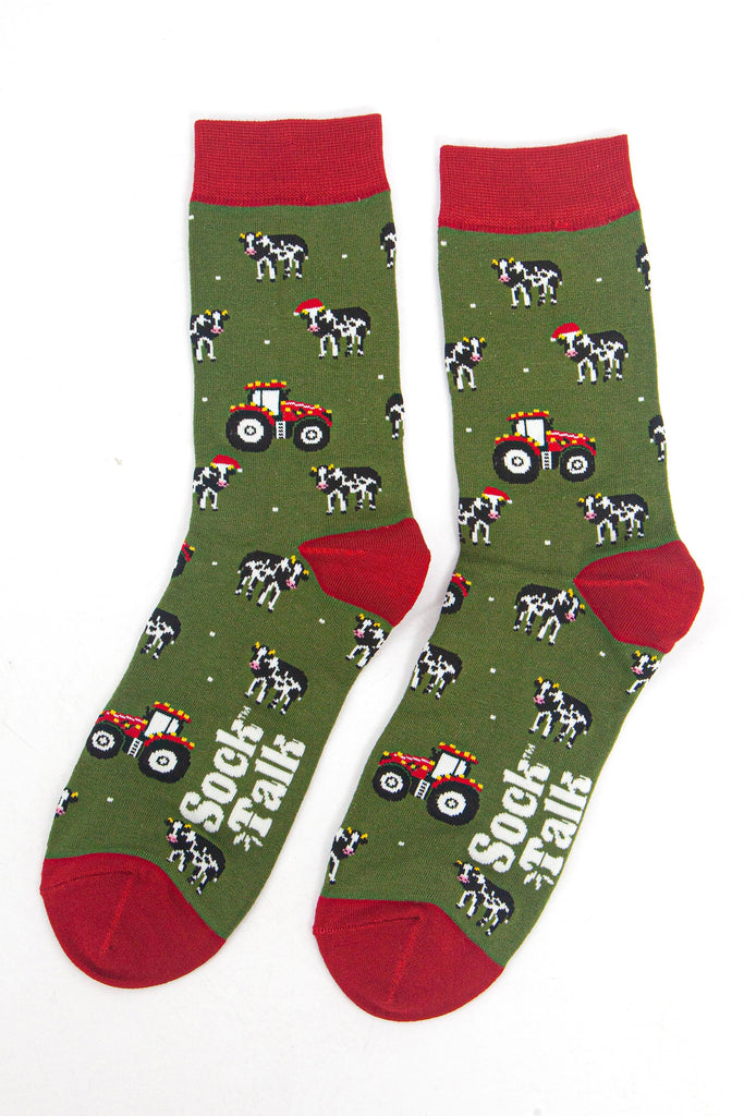 showing the tractor and cow socks laying flat, the cows are black and white with santa hats, the tractors are reed and have party lights on them. the toe, heel and cuff of the socks are red in contrast to the green body of the socks.