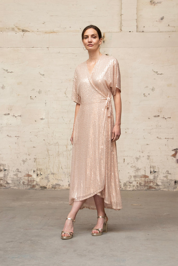 model wearing a gold sparkly sequin wrap dress accessorised with gold heeled sandals and earrings. the dress has an all over sparkle to it