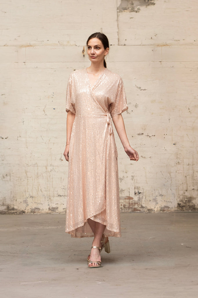 model wearing a gold sequin wrap dress, showing the waist tie feature and the dipped hem design of the piece