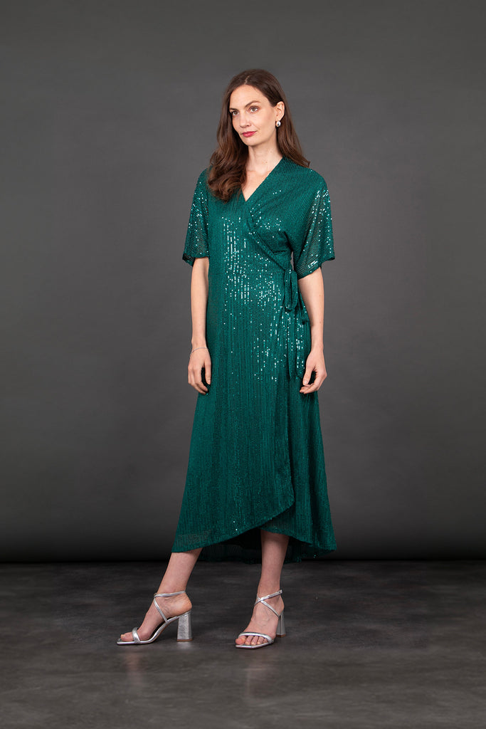 model wearing a dipped hem wrap dress in green with an all over sequin sparkle. the dress has a v-neck and waist tie