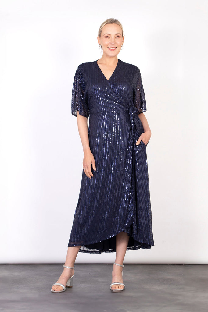 model wearing a navy blue midi length dipped hem sparkly sequinned wrap dress with a v-neck and waist tie fastening