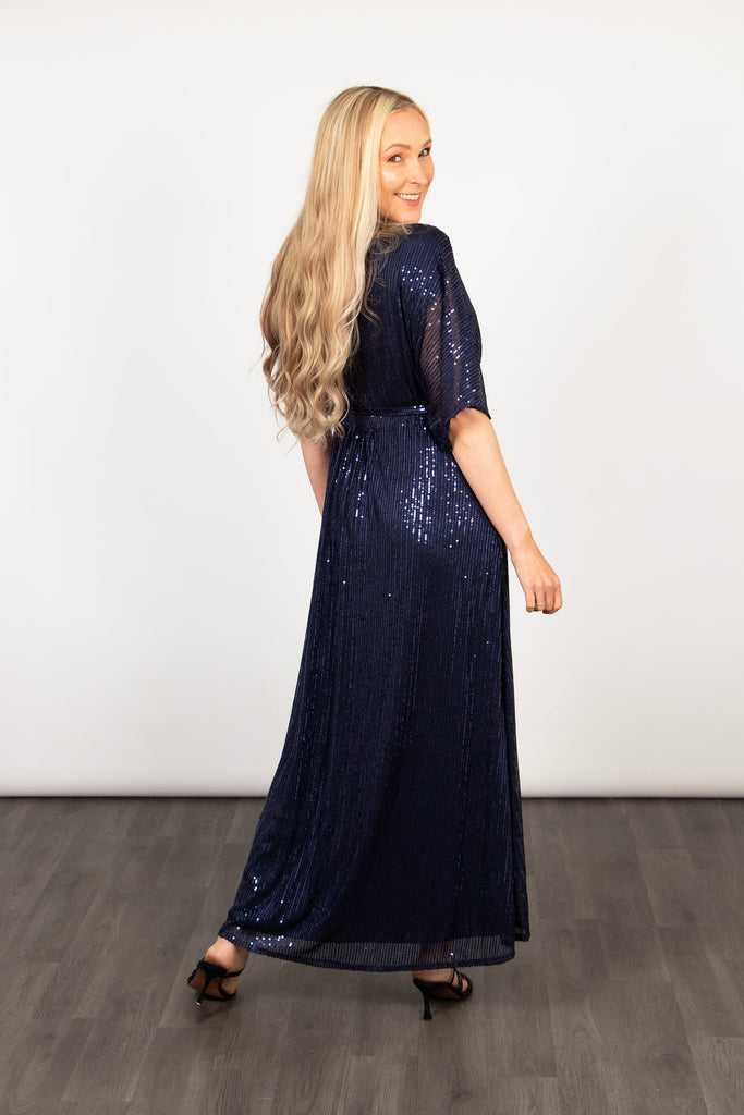 model showing the back of the blue sequin wrap dress, showing that the hem is longer at the back than at the front. it is maxi length at the back