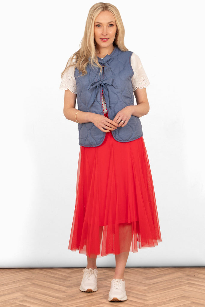 model wearing a red tulle skirt styled with a blue sleeveless gilet, white short sleeve blouse and white trainers showing a casual look