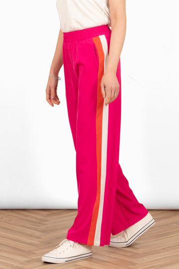 model wearing a pair of hot pink causal trousers with two vertical stripes running down the side of the leg. one stripe is orange the other is white.