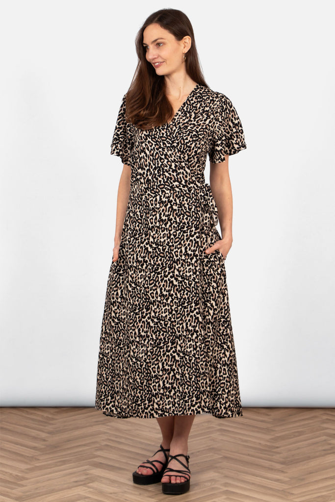 model wearing a neutral leopard print wrap dress. the waist tie is shown at tied in a bow at the side. the dress has a v-neck, short sleeves and side pockets.