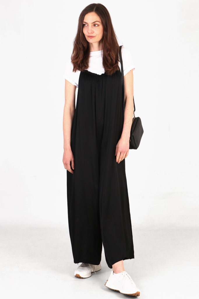 Model wearing black silky strappy jumpsuit with white cap sleeve tshirt underneath. Black leather bag on shoulder