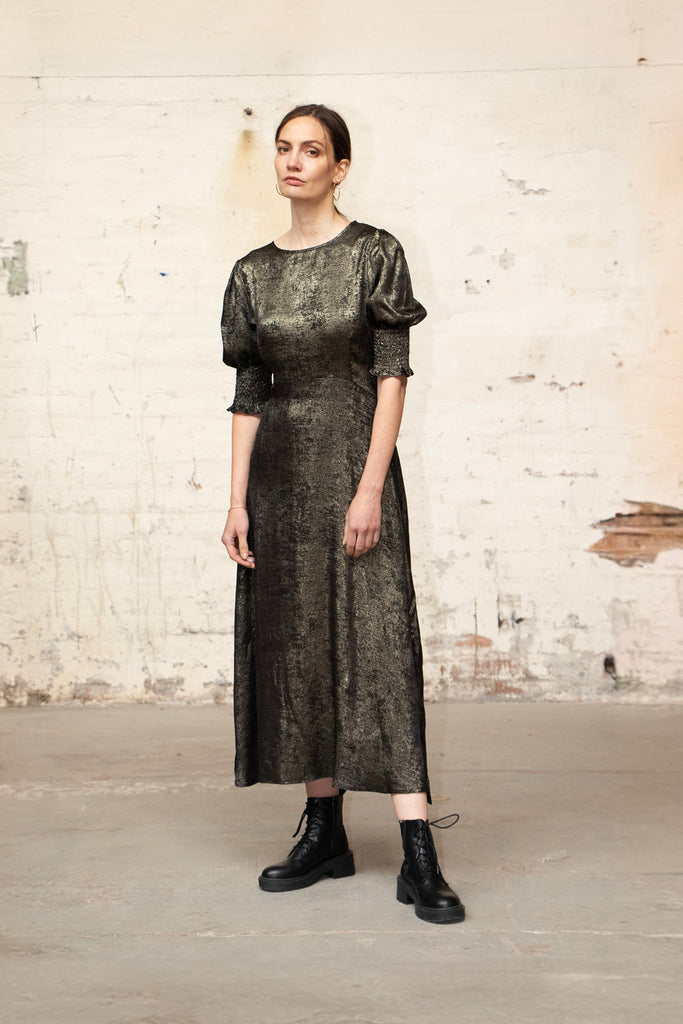 model wearing a metallic gold tea dress with a round neck and cuff sleeves. the dress is accessorised with black combat boots for an edgy look.