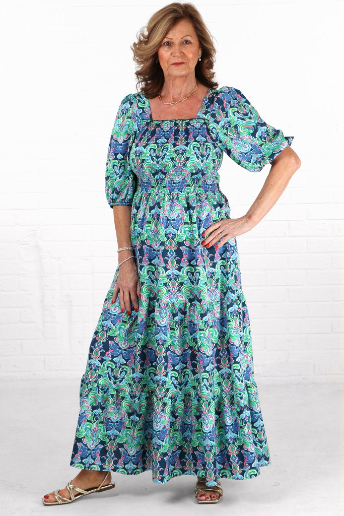 model wearing a blue tiered milkmaid maxi dress with short sleeves and an all over fleur de lis floral print
