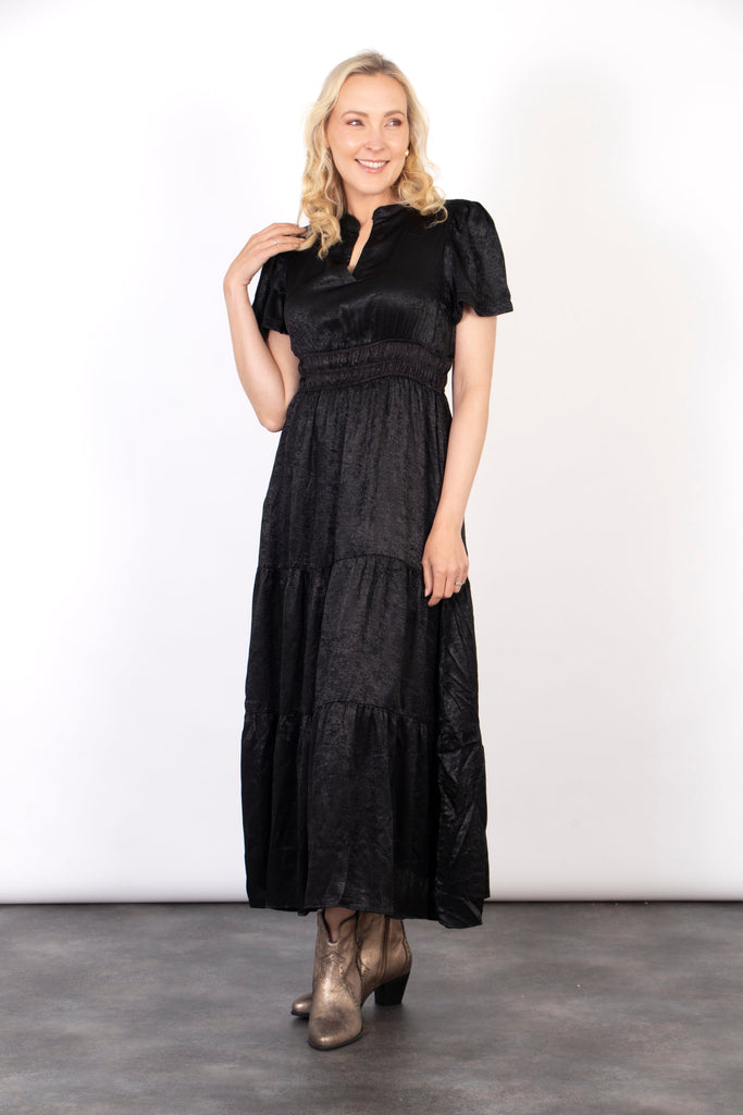 model wearing a short sleeve black tiered maxi dress with a metallic sheen all over. the dress has a cinched elasticated waist
