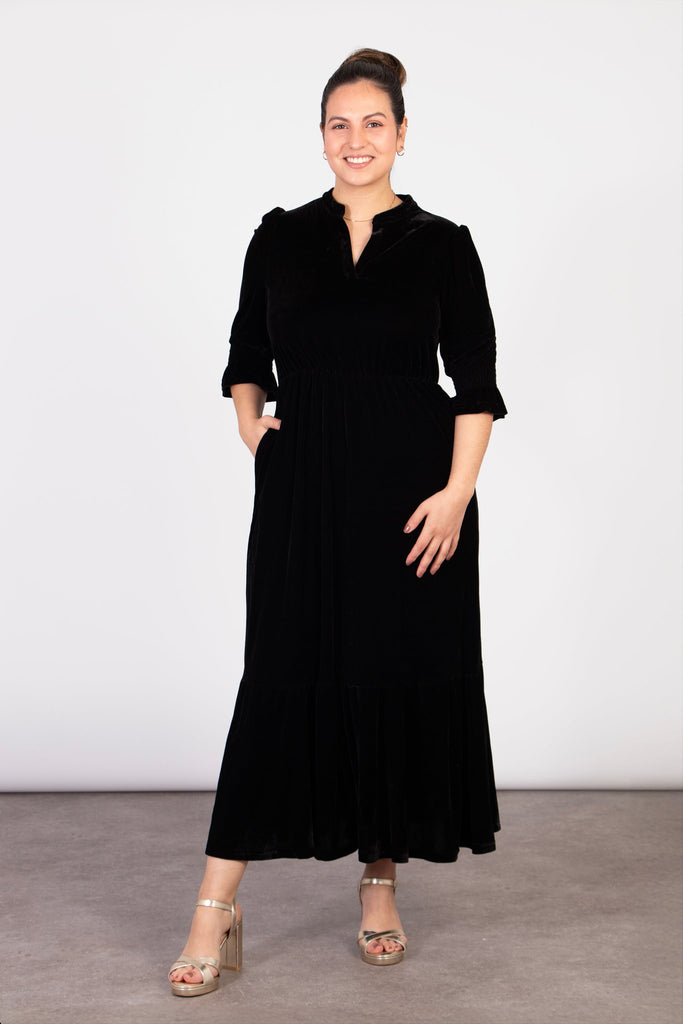 model wearing a midi length black velvet dress. the dress has pockets on the side of the design