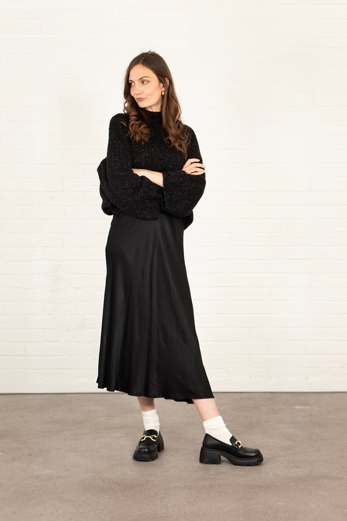 model wearing a plain black silky midi slip skirt, accessorised with a winter jumper and black shoes.
