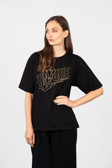 model wearing a black relaxed fitting t-shirt with a round neck and the word unique printed on the front in gold glitter, the letter i in unique is desigened to look like a lightning bolt