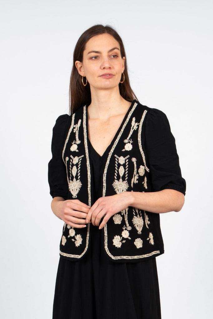 model wearing a black sleeveless lightweight waistcoat. the pattern on the waistcoat is an embroidered vintage floral design in ecru. 