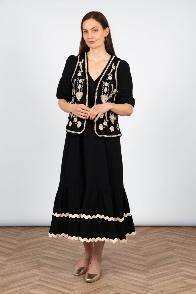 model wearing a black and ecru embroidered waistcoat styled worn over a black midi dress and paired with gold flat shoes.