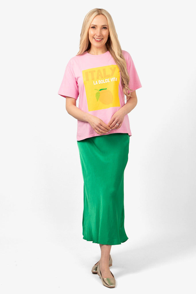 model wearing a pink cotton tshirt with a square yellow graphic logo on the front. the t-shirt is styled with a green midi slip skirt and gold flat pumps.