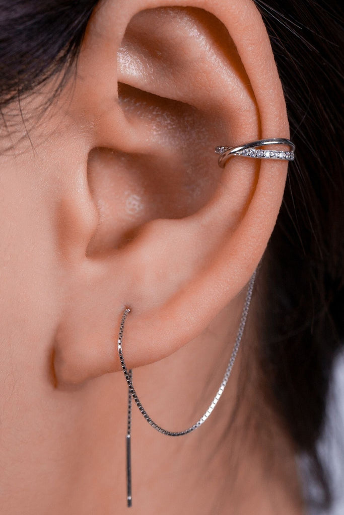 model wearing a sterling silver cross shaped clip on ear cuff  with a threader chain looping through an earlobe piercing. the cross shaped cuff has small white cubic zirconia crystals on them