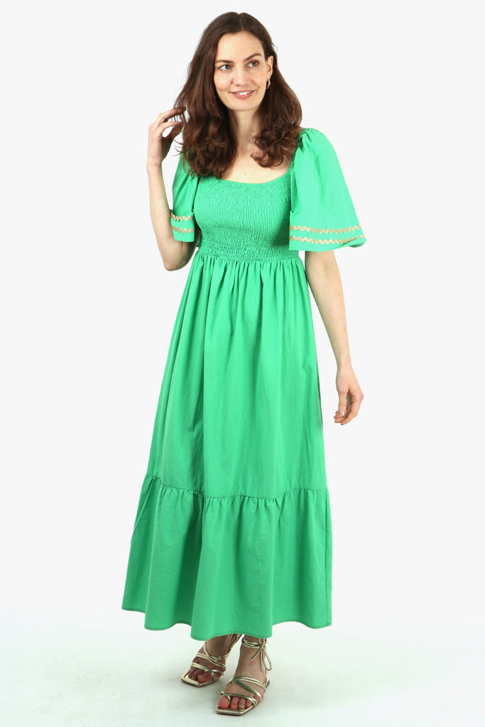 model wearing a green maxi length cotton tiered dress with shirred bust and short bell sleeves, the sleeves have gold wavy trimmed details